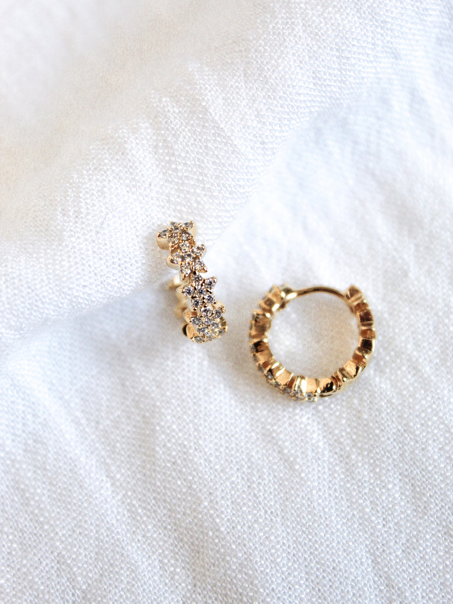 The Cloud Huggie earrings by Kinsey Designs Archives feature textured gold hoops with cubic zirconia accents, showcased on a white fabric background.