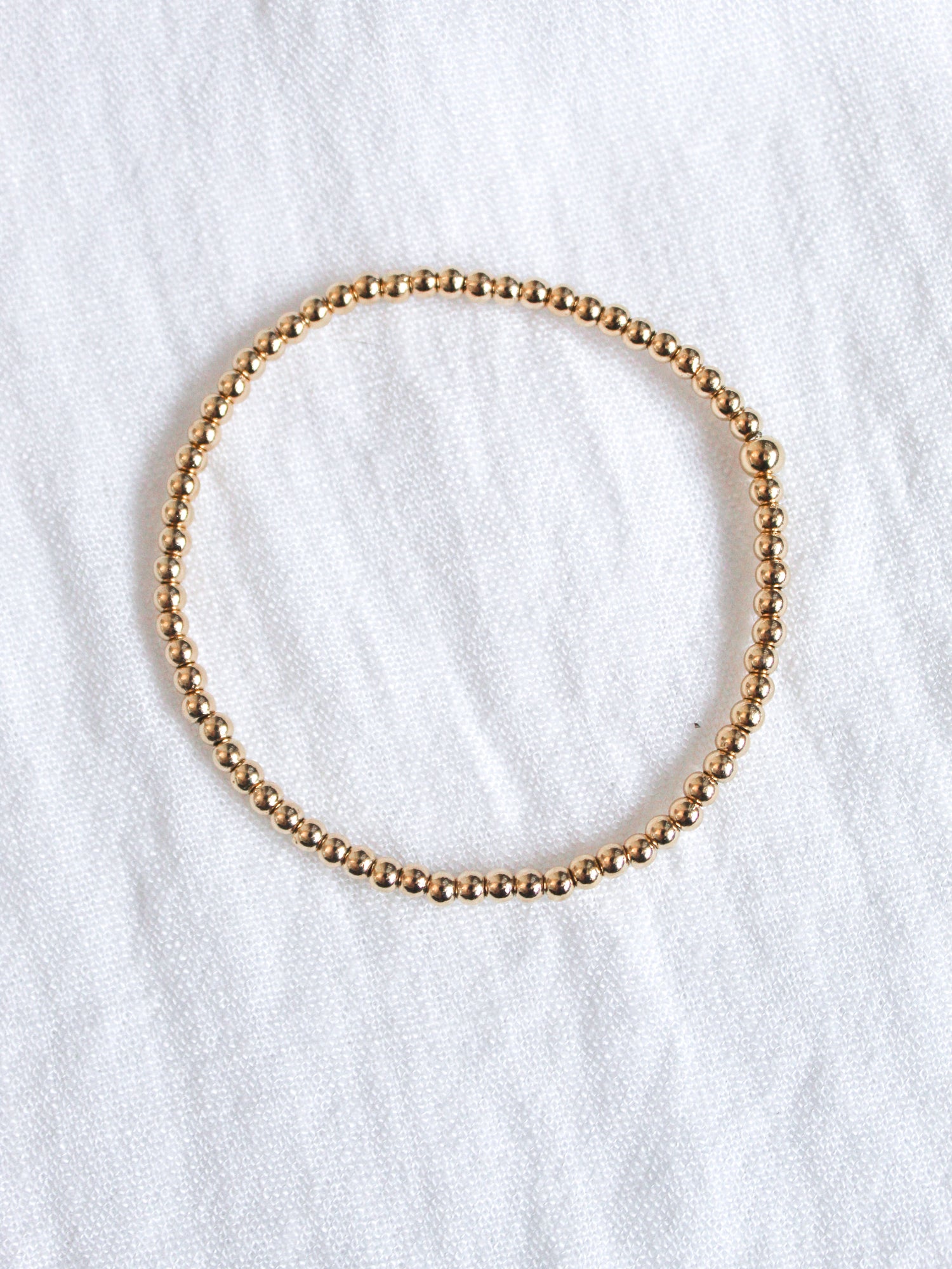 The Liam Stacking Bracelet by Kinsey Designs, shown in a circle on white textured fabric, features gold-filled beads and an adjustable stretch design for a comfortable fit. Available in multiple sizes.