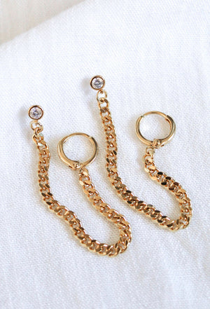 The Knox Double Earring by Kinsey Designs features clear cubic zirconia studs and delicate gold-plated chains, ideal for double piercings. Shown on a white fabric background.