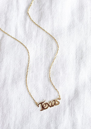 The Kinsey Designs Archives State of Mind Necklace features a gold filled pendant hand stamped with "Texas," displayed on a white textured fabric background.