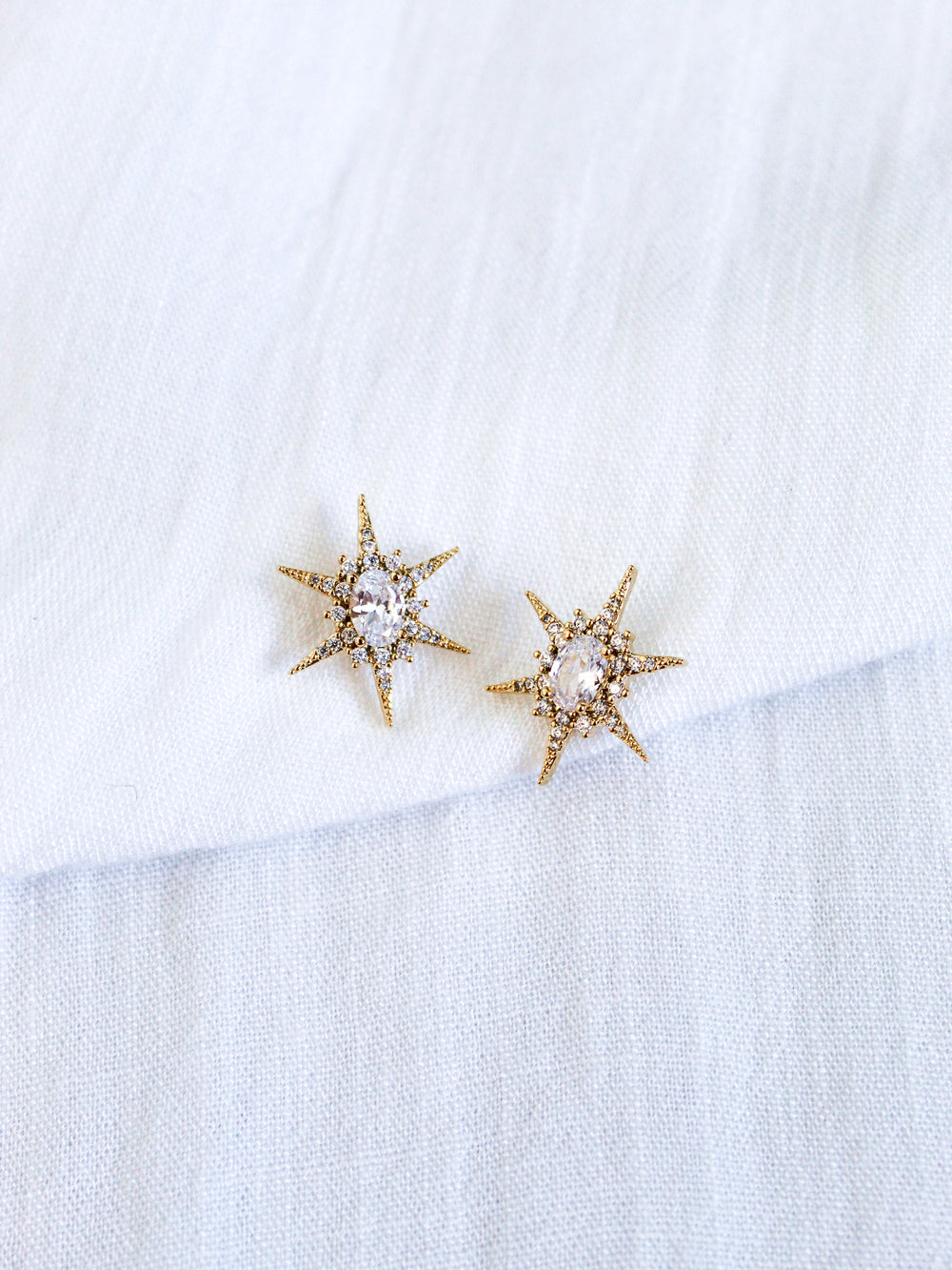 The Faye Stud by Kinsey Designs features gold star-shaped earrings with clear cubic zirconia gemstones in gold-filled metal, showcased on a white textured fabric background.