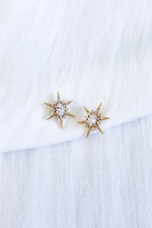The Faye Stud by Kinsey Designs features gold star-shaped earrings with clear cubic zirconia gemstones in gold-filled metal, showcased on a white textured fabric background.