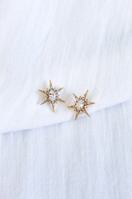 The Faye Stud by Kinsey Designs features gold star-shaped earrings with clear cubic zirconia gemstones in gold-filled metal, showcased on a white textured fabric background.
