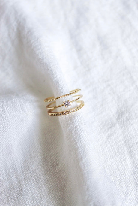 The Madi Ring by Kinsey Designs Archives is an adjustable gold plated ring featuring three bands and a small diamond, shown on a soft white fabric background.
