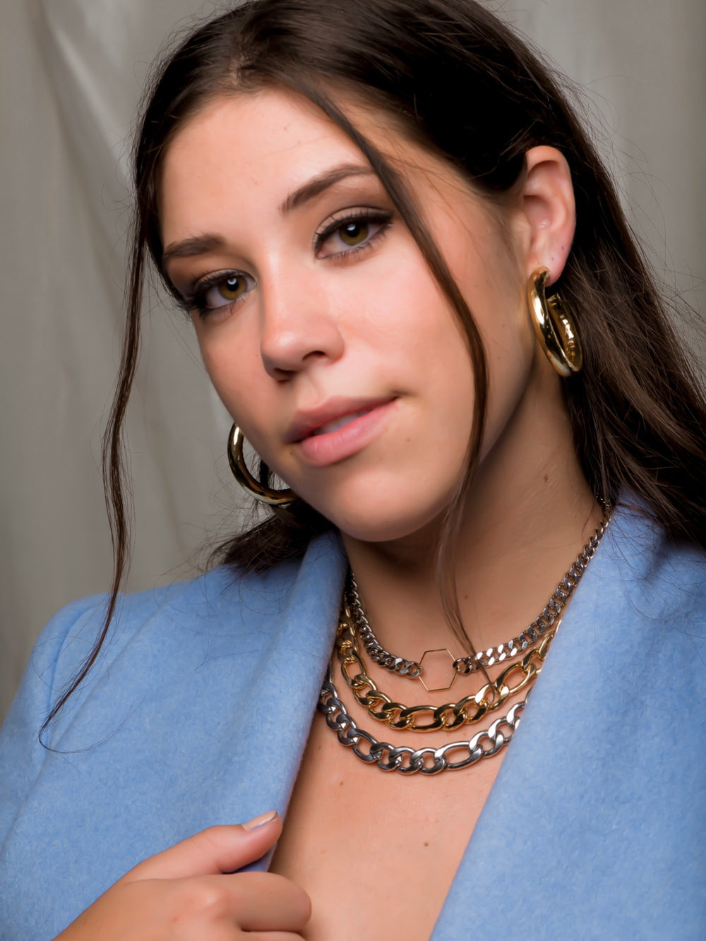 A woman with long brown hair wears Kinsey Designs' Jasmine Hoops, layered necklaces, and a blue jacket, showcasing statement jewelry indoors.