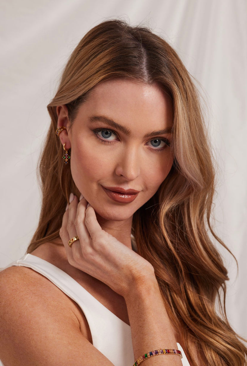 Woman with long wavy hair, wearing the Luna Ring - Multi by Kinsey Designs Archives, dressed in a white top and smiling softly while touching her neck.