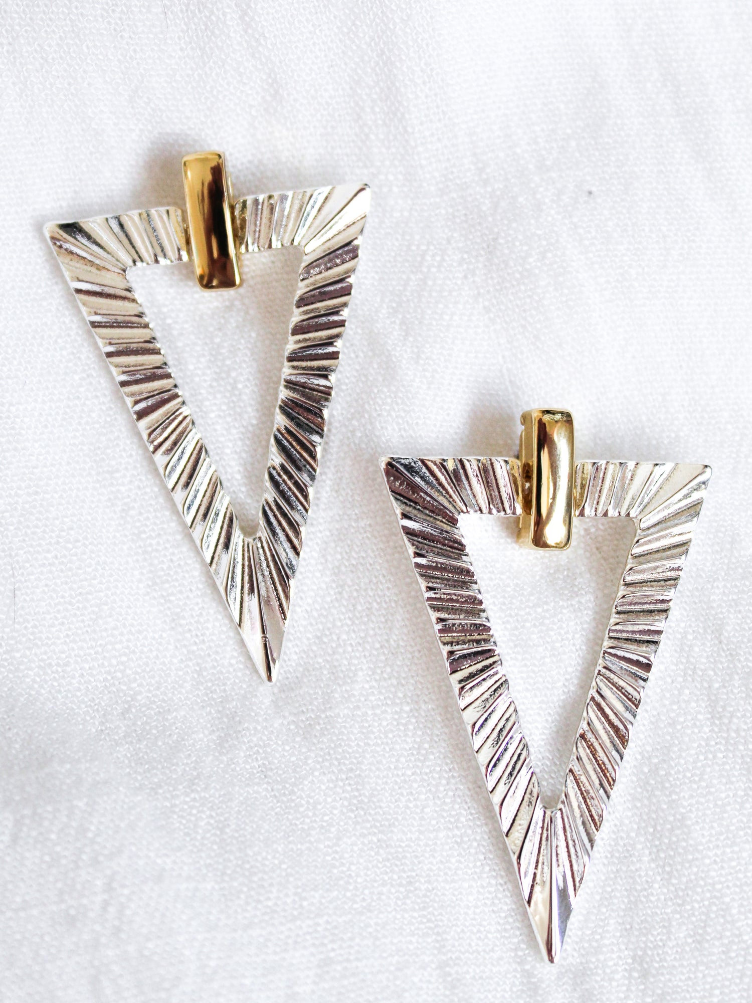 The Colin Earring by Kinsey Designs features silver triangular gold-filled earrings with textured lines and gold-colored top accents, displayed on white fabric. Perfect for a chic look—shop the sample sale now!.