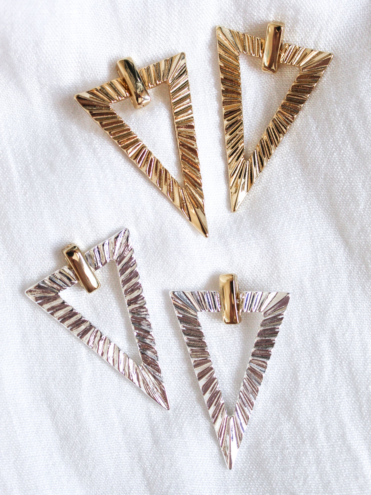 Four textured, triangular metal Colin Earrings by Kinsey Designs—two gold, two silver—styled on a white fabric background. Ideal for collecting or gifting at our exclusive sample sale.