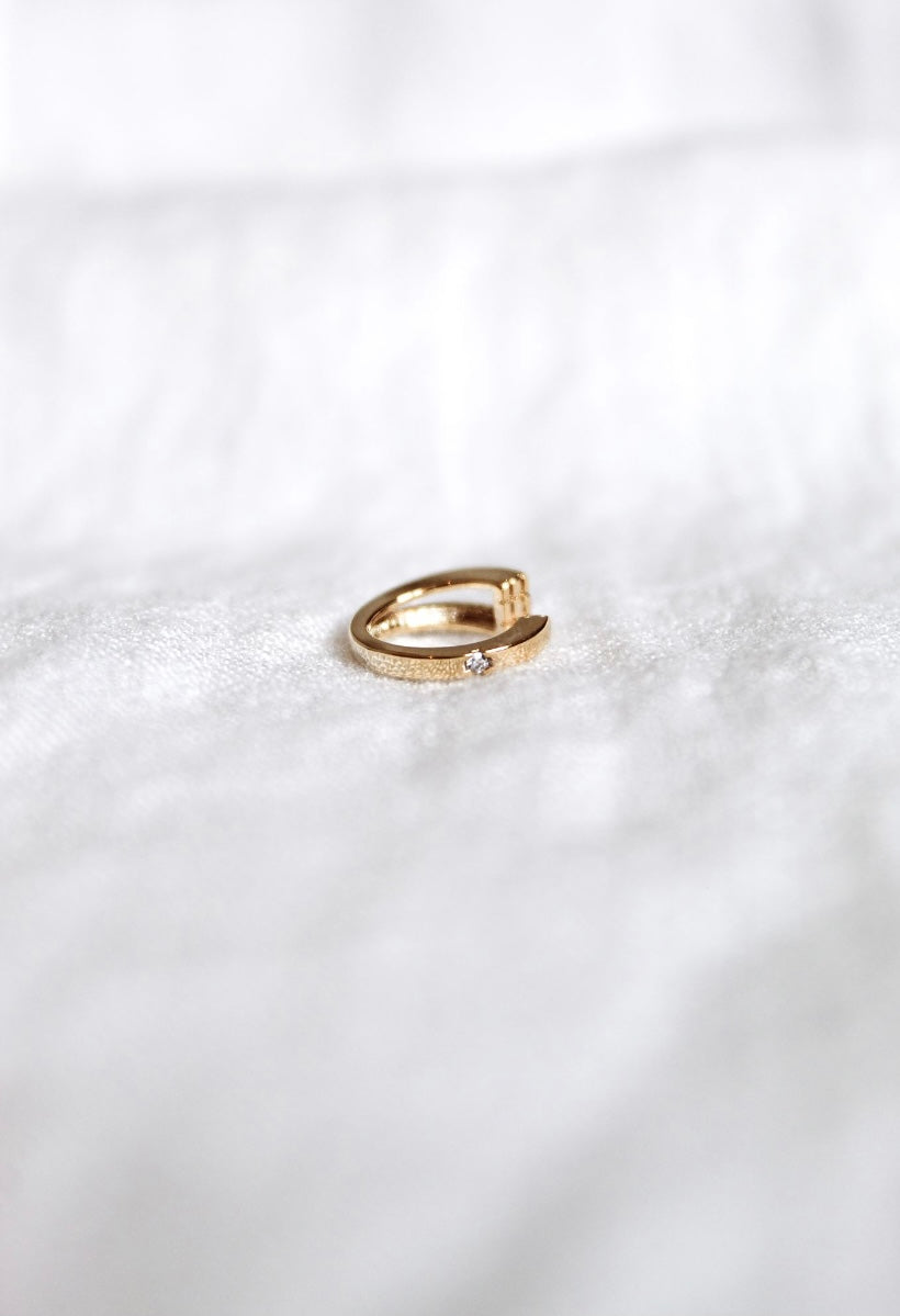 The Leni Ear Cuff by Kinsey Designs Archives, featuring gold plating and a small cubic zirconia, rests on white textured fabric, adding subtle elegance to the jewelry display.