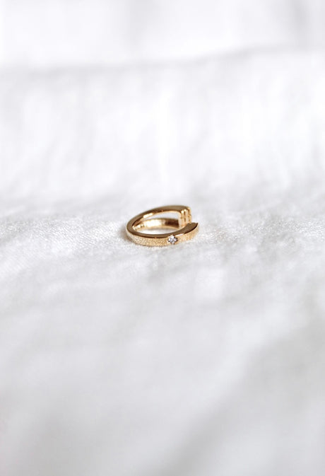 The Leni Ear Cuff by Kinsey Designs Archives, featuring gold plating and a small cubic zirconia, rests on white textured fabric, adding subtle elegance to the jewelry display.