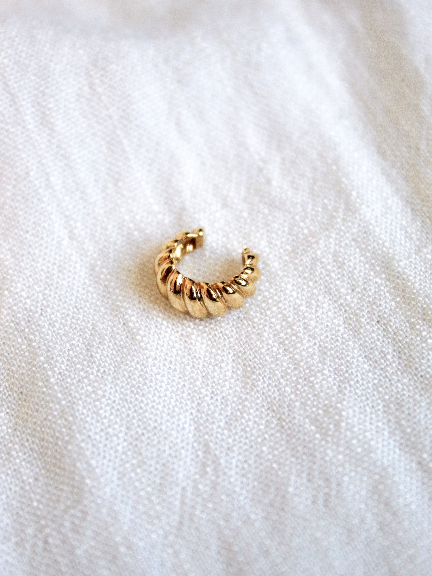 The Metz Cuff by Kinsey Designs Archives, a gold-plated textured ear cuff, rests on a soft white fabric surface.