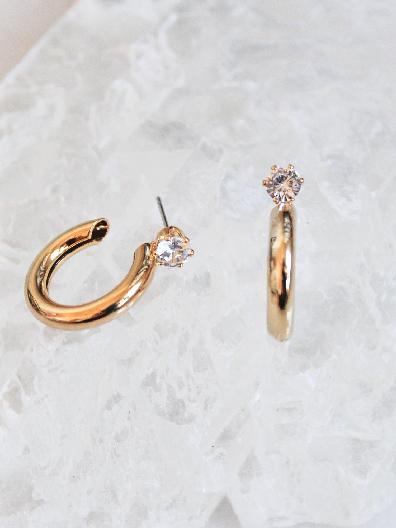 The Hugo earrings by Kinsey Designs Archives are two 1-inch wide gold-plated hoops with clear gemstones, shown on a textured white surface.
