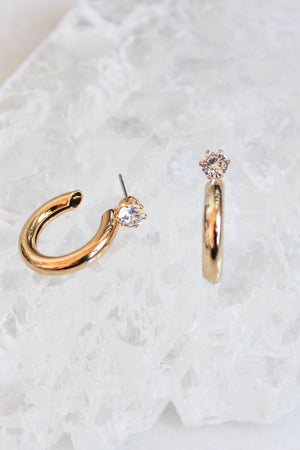 The Hugo earrings by Kinsey Designs Archives are two 1-inch wide gold-plated hoops with clear gemstones, shown on a textured white surface.