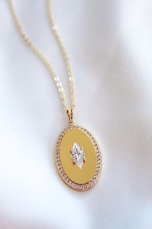 The Fique Necklace by Kinsey Designs features a gold-plated 32-inch chain with a central cubic zirconia pendant encircled by sparkling stones, beautifully displayed on white fabric—perfect for elegant layering.