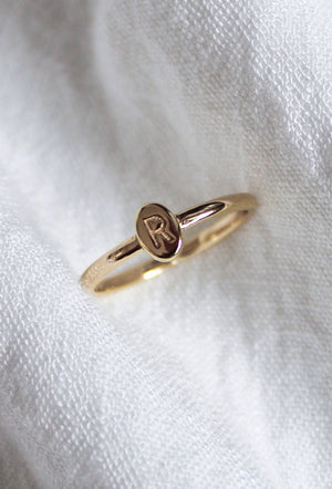 The Kinsey Initial Ring by Kinsey Designs features an engraved letter "R" and rests elegantly on white textured fabric, making it perfect as personalized jewelry.