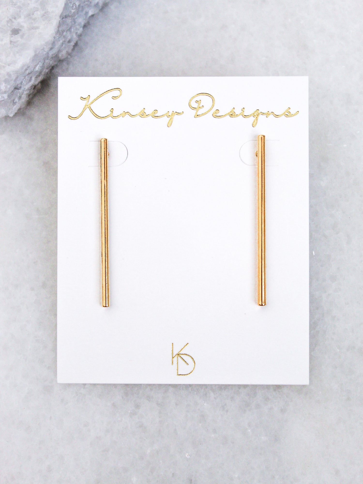 Walker gold bar earrings by Kinsey Designs Archives, featuring a surgical steel post, shown on a white branded card against a light marble background.