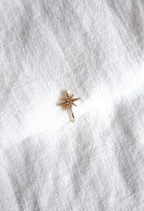 The Burst Ear Cuff by Kinsey Designs Archives is a gold plated, star-shaped ear cuff adorned with sparkling cubic zirconia, shown on white textured fabric.