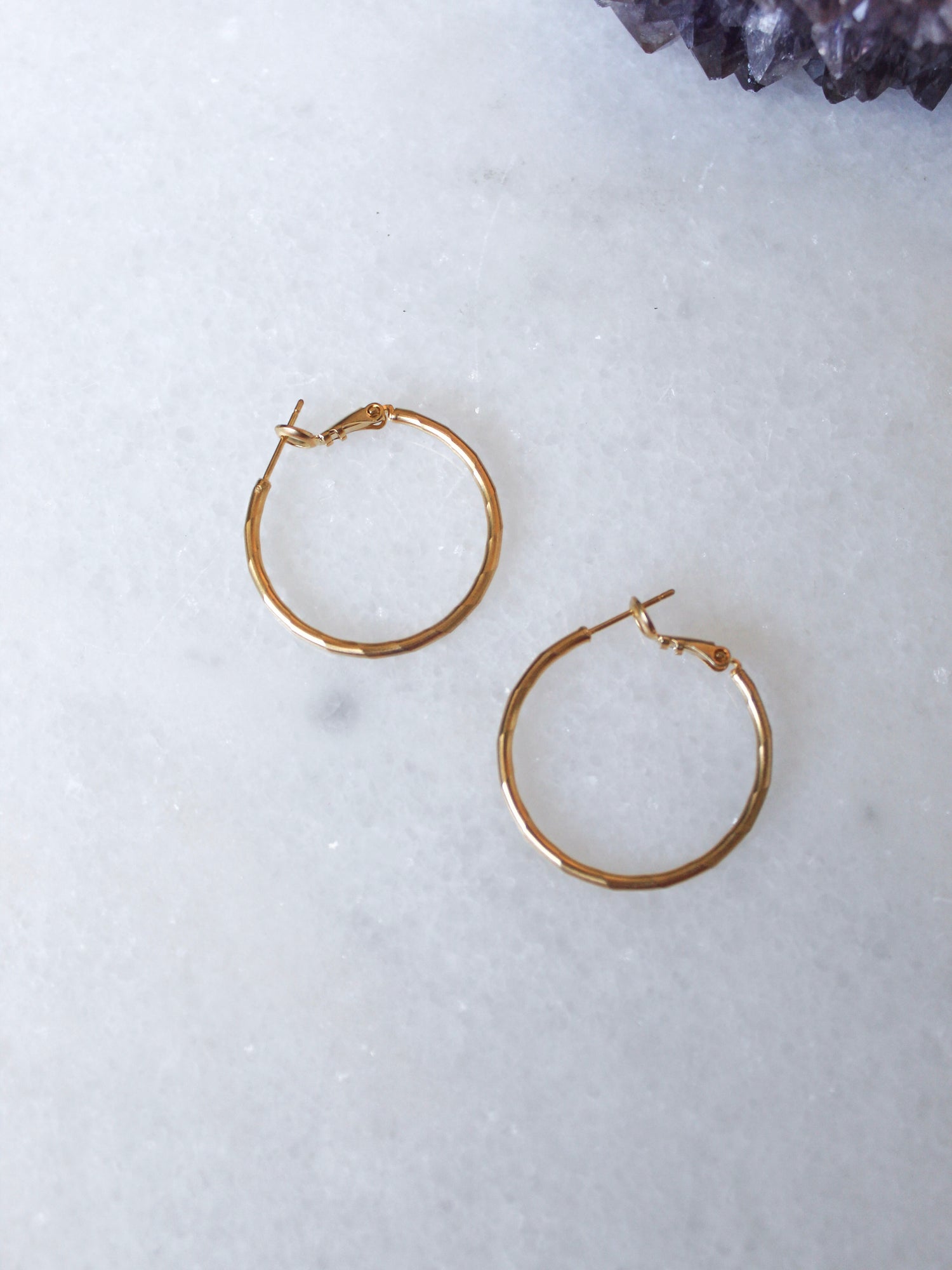Two small Lexie 18k gold plated hoop earrings by Kinsey Designs Archives rest on a white marble surface, with a purple crystal in the corner.