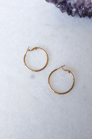 Two small Lexie 18k gold plated hoop earrings by Kinsey Designs Archives rest on a white marble surface, with a purple crystal in the corner.