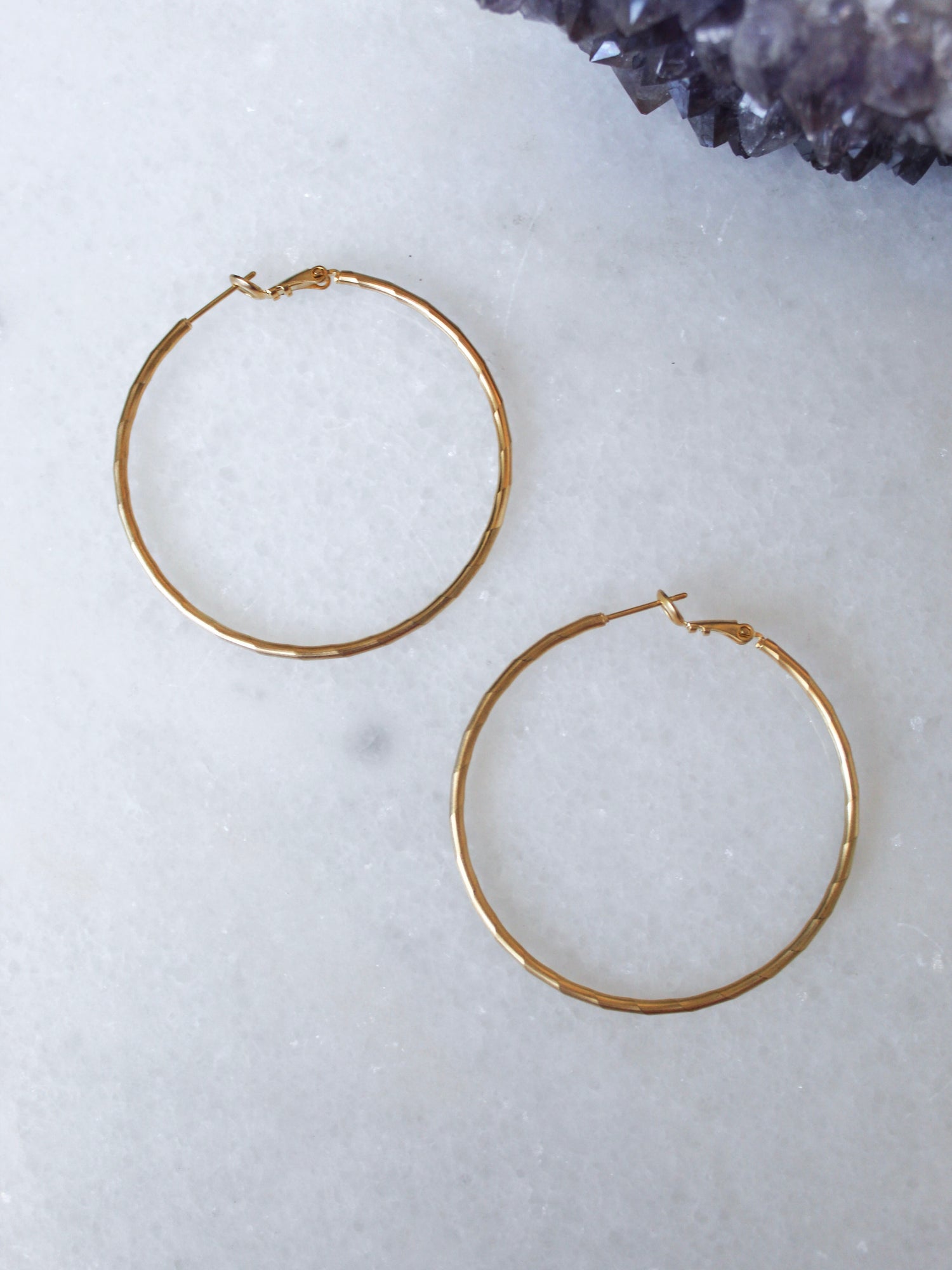 The Lexie earrings by Kinsey Designs Archives—one large 2” and one small 1”, both 18k gold plated hoops—are displayed on a white marble surface with purple crystals in the top right corner.