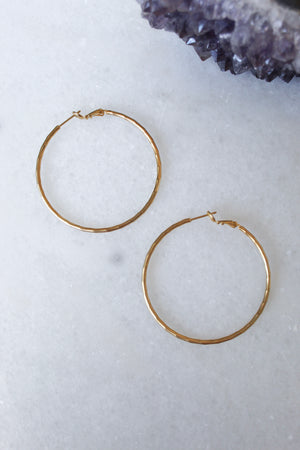 The Lexie earrings by Kinsey Designs Archives—one large 2” and one small 1”, both 18k gold plated hoops—are displayed on a white marble surface with purple crystals in the top right corner.