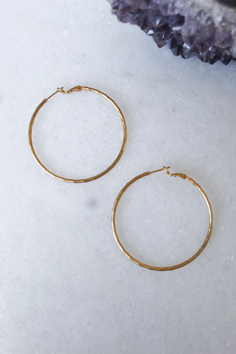 The Lexie earrings by Kinsey Designs Archives—one large 2” and one small 1”, both 18k gold plated hoops—are displayed on a white marble surface with purple crystals in the top right corner.