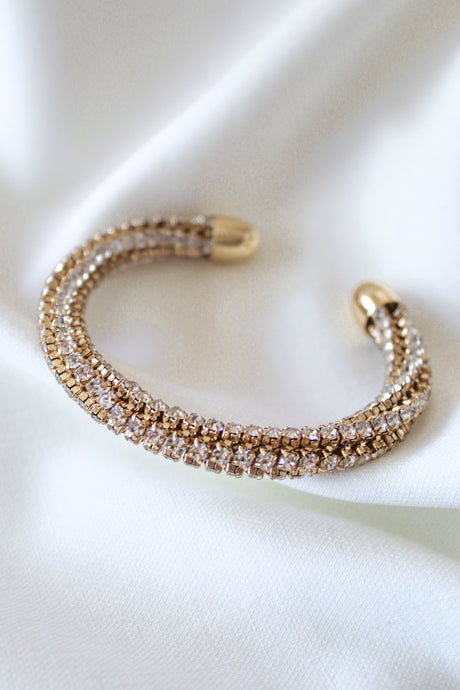 The Smog Cuff by Kinsey Designs Archives features 16k gold-plated brass and two rows of sparkling cubic zirconia crystals, elegantly showcased against a white fabric backdrop.